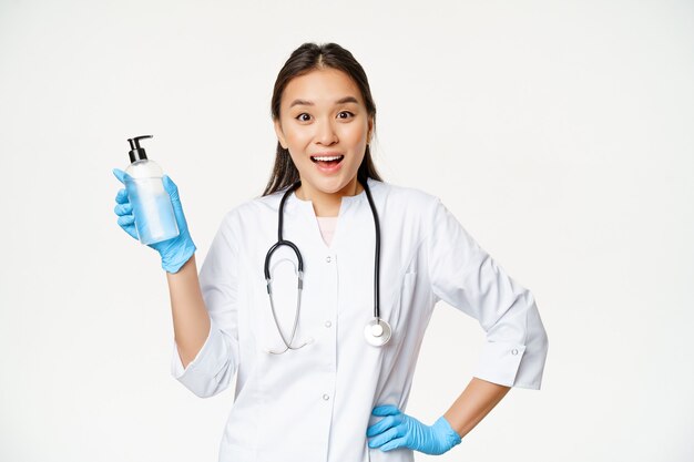 Doctora entusiasta en uniforme, mostrando jabón de manos, desinfectante, prevención de covid-19, de pie sobre fondo blanco.