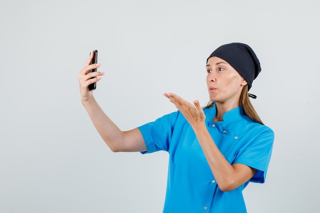 Doctora enviando beso de aire mientras toma selfie en uniforme azul, sombrero negro