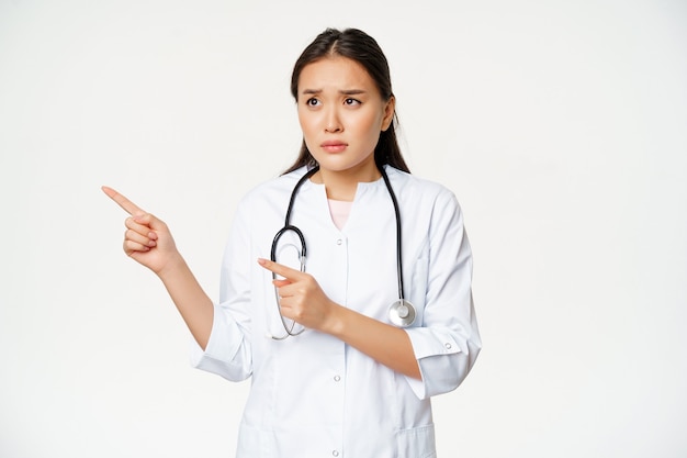 Doctora escéptica, enfermera preocupada apuntando y mirando a la izquierda con expresión de cara preocupada, de pie en bata médica blanca sobre fondo de estudio.