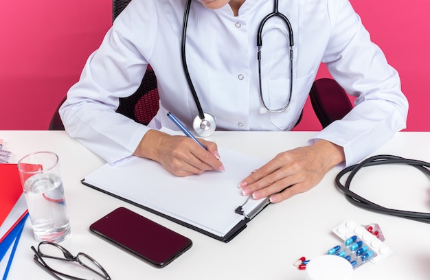 Doctora eslava adulta complacida en bata médica con estetoscopio sentado en el escritorio con herramientas de oficina escribiendo en el portapapeles aislado en la pared rosa con espacio de copia