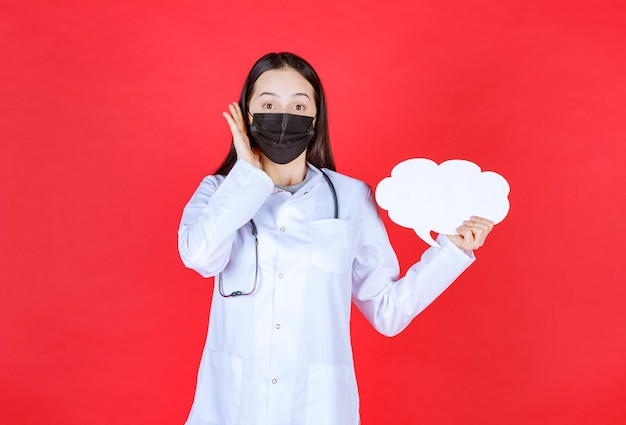 Doctora con estetoscopio y máscara negra sosteniendo un escritorio de información en blanco con forma de nube y abriendo la oreja para escuchar bien.