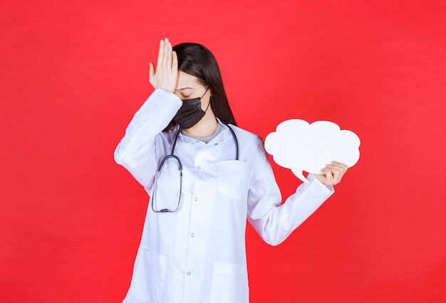 Doctora con estetoscopio y máscara negra sosteniendo un escritorio de información en blanco con forma de nube y sosteniendo la cabeza.