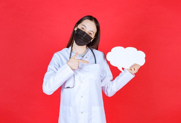 Doctora con estetoscopio y máscara negra sosteniendo un escritorio de información en blanco con forma de nube.