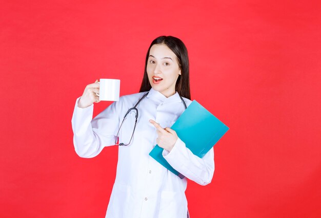 Doctora con estetoscopio sosteniendo una taza de bebida y una carpeta de historial del paciente.