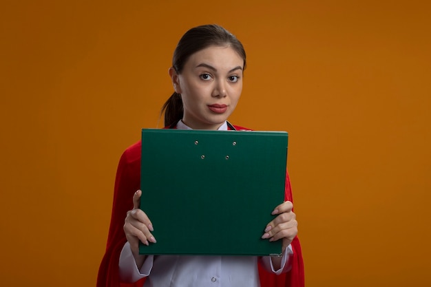 Doctora con estetoscopio en uniforme médico blanco y capa roja de superhéroe sosteniendo una carpeta frente a ella mirando al frente con cara seria de pie sobre la pared naranja