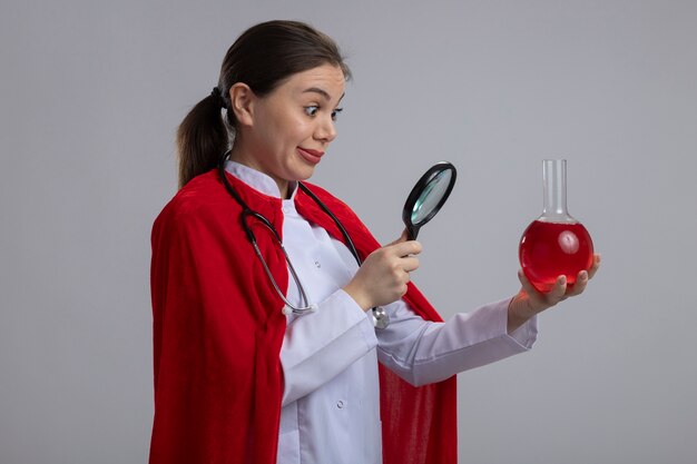 Doctora con estetoscopio en uniforme médico blanco y capa de superhéroe roja sosteniendo el matraz con líquido rojo mirándolo a través de una lupa confundido de pie sobre una pared blanca