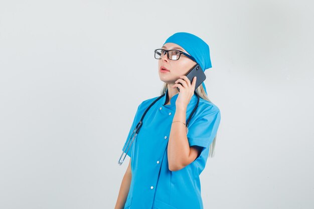 Doctora hablando por teléfono inteligente y mirando hacia arriba en uniforme azul