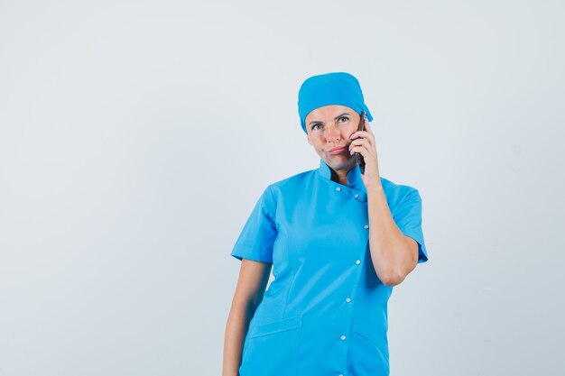 Doctora hablando por teléfono móvil en uniforme azul y mirando vacilante, vista frontal.