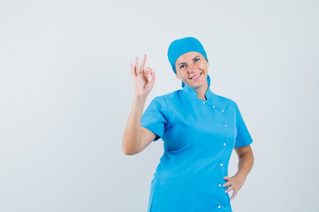Doctora haciendo un gesto aceptable en uniforme azul y mirando confiado, vista frontal.