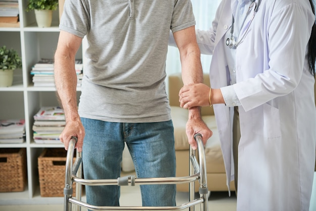 Doctora irreconocible ayudando a paciente masculino a caminar con bastidor