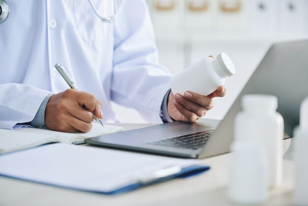 Doctora irreconocible con laptop sosteniendo medicamentos y escribiendo recetas