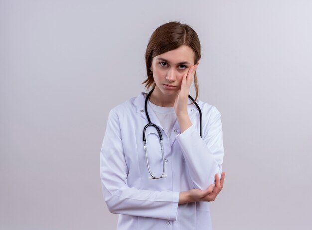 Doctora joven aburrida vistiendo bata médica y estetoscopio y poniendo la mano en la mejilla con espacio de copia