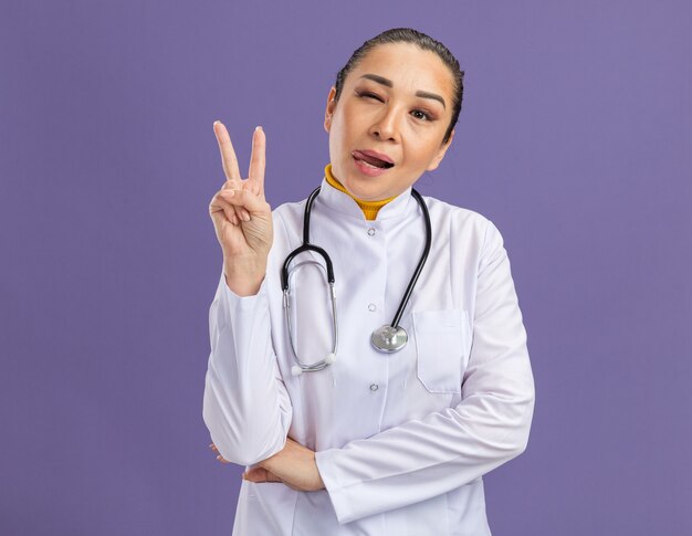 Doctora joven alegre en bata blanca de medicina con estetoscopio alrededor del cuello que muestra el signo v sacando la lengua guiñando un ojo feliz y positivo de pie sobre la pared púrpura