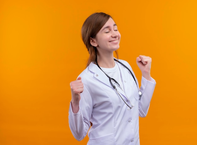 Doctora joven alegre con bata médica y un estetoscopio con los puños apretados y los ojos cerrados en el espacio naranja aislado con espacio de copia