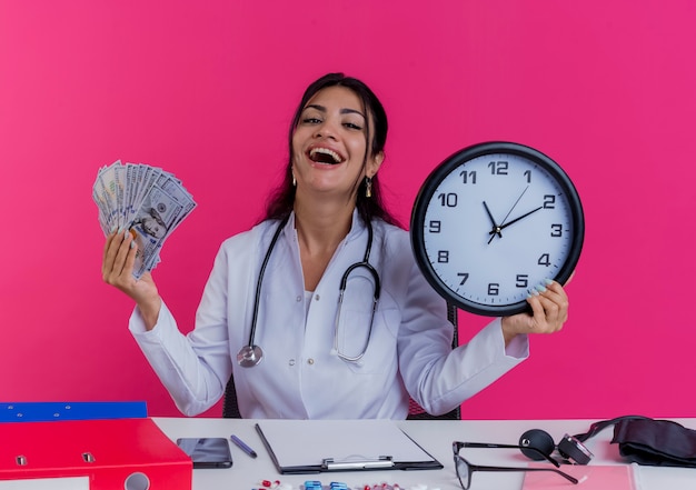 Doctora joven alegre con bata médica y un estetoscopio sentado en el escritorio con herramientas médicas con dinero y reloj aislado en la pared rosa