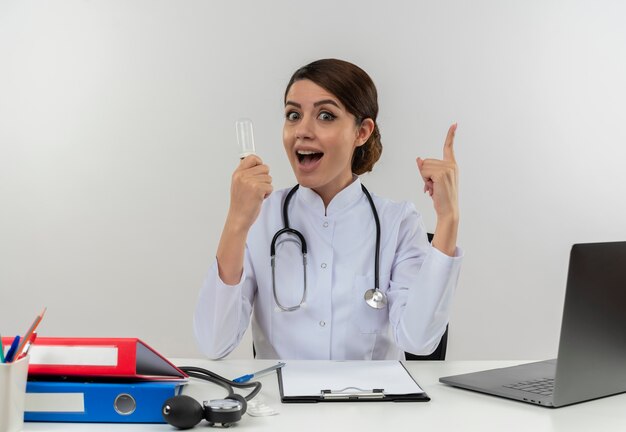 Doctora joven alegre con bata médica con estetoscopio sentado en el escritorio de trabajo en la computadora con herramientas médicas sosteniendo la bombilla con el pulgar hacia arriba en la pared blanca aislada con espacio de copia