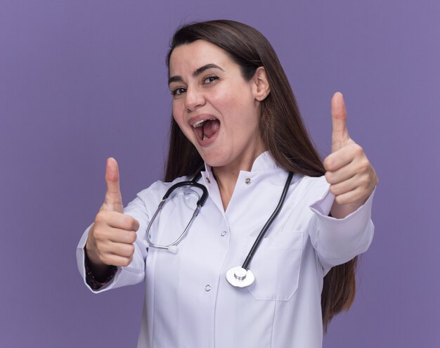 Doctora joven alegre vistiendo bata médica con estetoscopio pulgar hacia arriba en púrpura