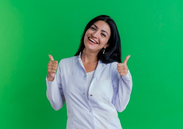 Doctora joven alegre vistiendo bata médica mostrando los pulgares para arriba aislado en la pared verde con espacio de copia