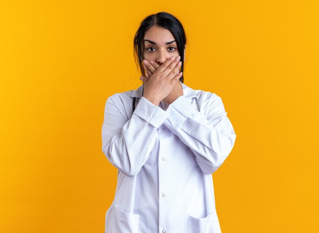 Doctora joven asustada vistiendo bata médica con estetoscopio cubrió la boca con las manos aisladas sobre fondo amarillo