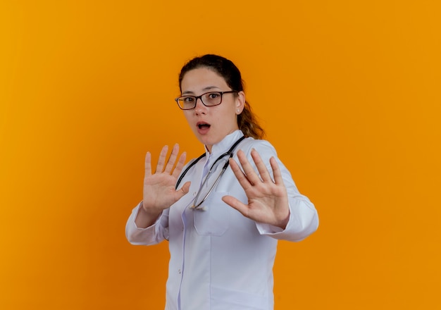 Doctora joven asustada vistiendo bata médica y estetoscopio con gafas mostrando gesto de parada aislado