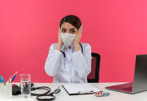 Doctora joven asustada vistiendo bata médica con estetoscopio en máscara médica sentado en el escritorio de trabajo en la computadora con herramientas médicas poniendo las manos en la cabeza con espacio de copia