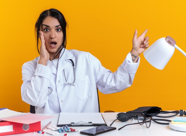 Doctora joven asustada vistiendo bata médica con estetoscopio se sienta a la mesa con puntos de herramientas médicas en el lado aislado sobre fondo amarillo con espacio de copia