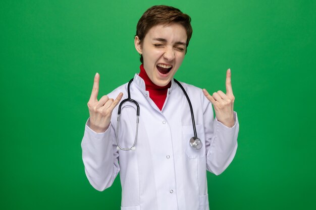 Doctora joven en bata blanca con estetoscopio gritando feliz y emocionado mostrando el símbolo de la roca