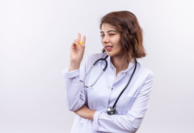 Doctora joven en bata blanca con estetoscopio guiñando un ojo mostrando el signo de la victoria con la lengua fuera de la pared blanca