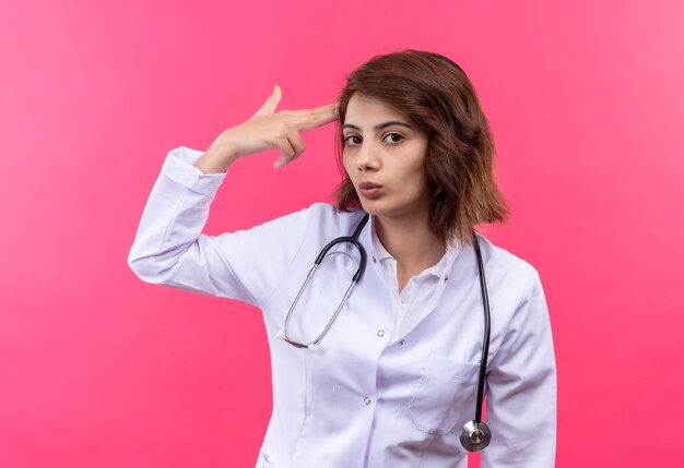 Doctora joven en bata blanca con estetoscopio haciendo pistola de dedo o gesto de pistola cerca de la sien de pie sobre la pared rosa
