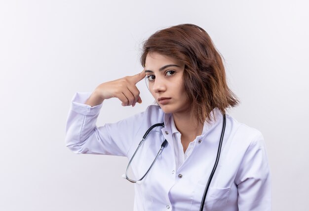 Doctora joven en bata blanca con estetoscopio mirando a cámara con expresión seria apuntando su sien