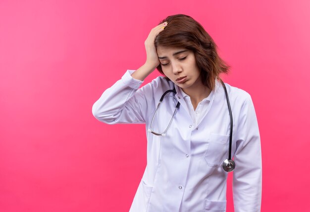 Doctora joven en bata blanca con estetoscopio mirando cansado y con exceso de trabajo con las manos en la cabeza con los ojos cerrados