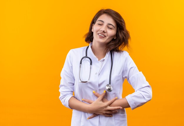 Doctora joven en bata blanca con estetoscopio con los ojos cerrados tocando su vientre sonriendo de pie sobre la pared naranja