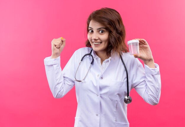 Doctora joven en bata blanca con estetoscopio sosteniendo el frasco de prueba apretando el puño feliz y salido