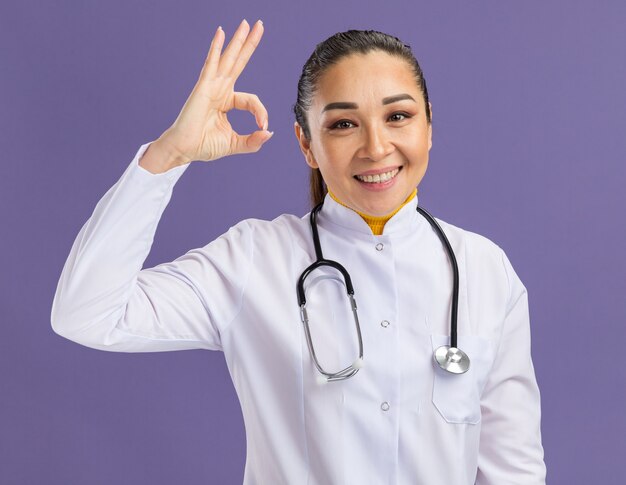 Doctora joven en bata blanca de medicina con estetoscopio alrededor del cuello con una sonrisa en la cara haciendo bien firmar de pie sobre la pared púrpura