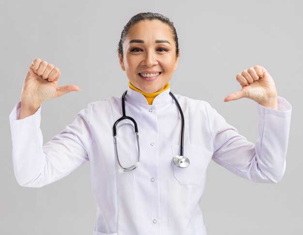 Doctora joven en bata médica blanca con estetoscopio alrededor del cuello sonriendo confiada apuntando a sí misma de pie sobre la pared blanca