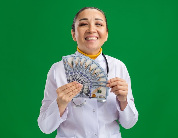 Doctora joven en bata médica blanca con estetoscopio alrededor del cuello sosteniendo dinero en efectivo con cara feliz de pie sobre la pared verde