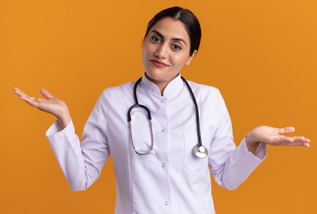 Doctora joven en bata médica con estetoscopio alrededor de su cuello mirando al frente confundido extendiendo los brazos hacia los lados sin respuesta de pie sobre la pared naranja