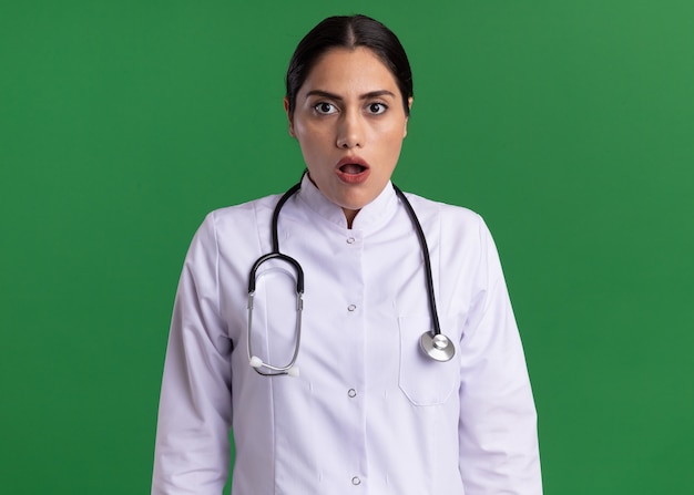 Doctora joven en bata médica con estetoscopio alrededor de su cuello mirando al frente confundido y preocupado de pie sobre la pared verde