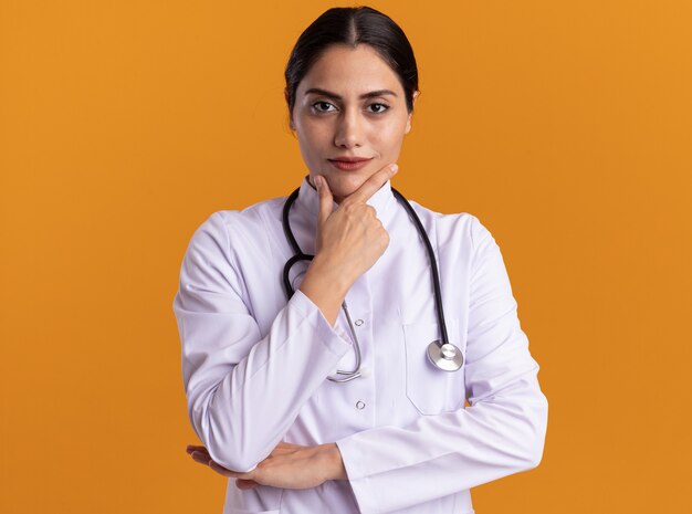 Doctora joven en bata médica con estetoscopio alrededor de su cuello mirando al frente con expresión seria y segura de pie sobre la pared naranja