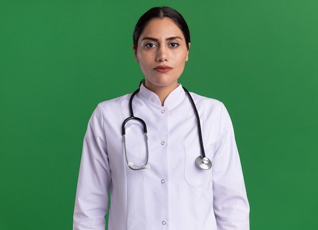 Doctora joven en bata médica con estetoscopio alrededor de su cuello mirando al frente con seria expresión de confianza de pie sobre la pared verde