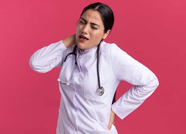 Doctora joven en bata médica con estetoscopio buscando malestar tocando su cuello sintiendo dolor de pie sobre la pared rosa
