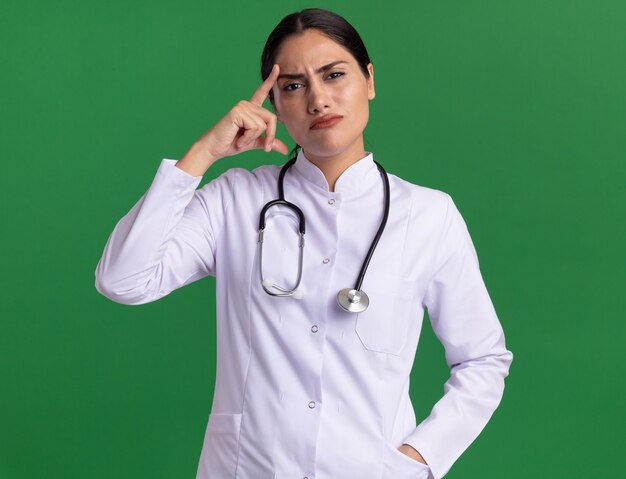 Doctora joven en bata médica con estetoscopio mirando confundido apuntando con el dedo índice a su sien de pie sobre la pared verde