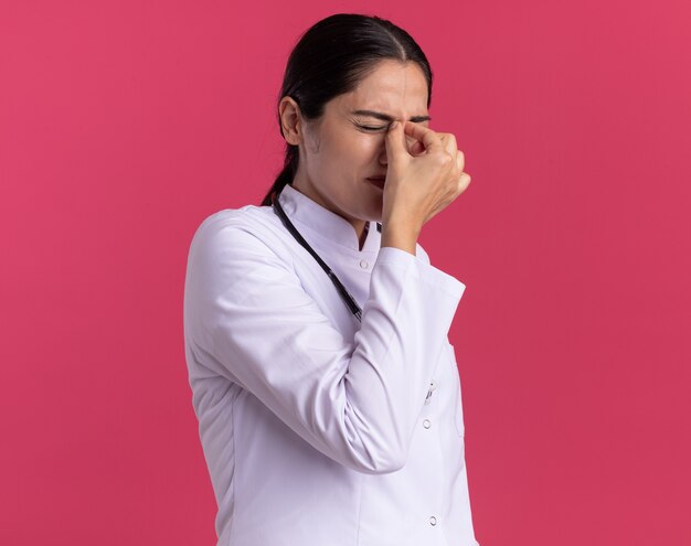 Doctora joven en bata médica con estetoscopio tocando la nariz entre los ojos cerrados con expresión molesta de pie sobre la pared rosa