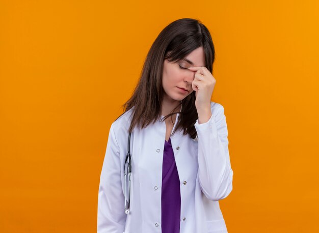 Doctora joven cansada en bata médica con estetoscopio pone la mano en la cara sobre fondo naranja aislado con espacio de copia