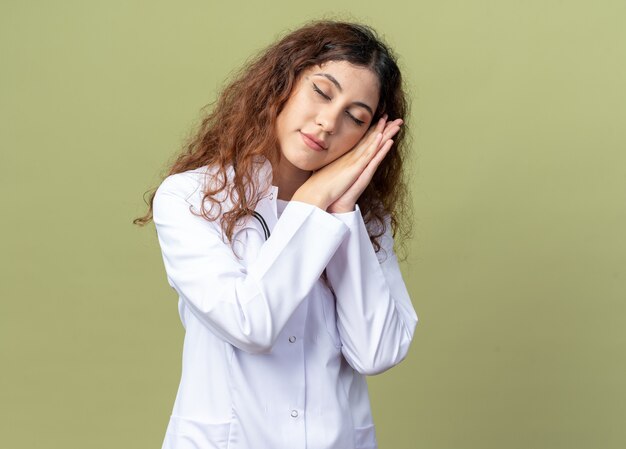 Doctora joven cansada vistiendo bata médica y un estetoscopio haciendo gesto de sueño con los ojos cerrados aislados en la pared verde oliva con espacio de copia
