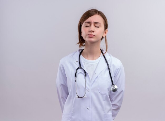 Doctora joven cansada vistiendo bata médica y estetoscopio con los ojos cerrados en la pared blanca aislada con espacio de copia