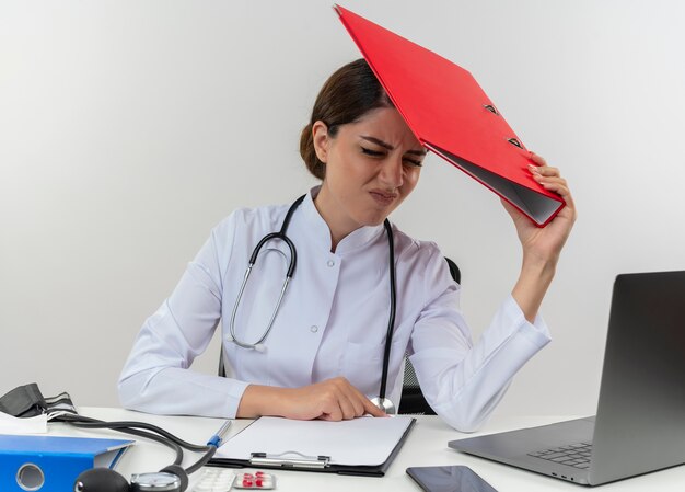 Doctora joven cansada vistiendo bata médica con estetoscopio sentado en el escritorio de trabajo en la computadora con herramientas médicas poniendo carpeta en la cabeza con espacio de copia