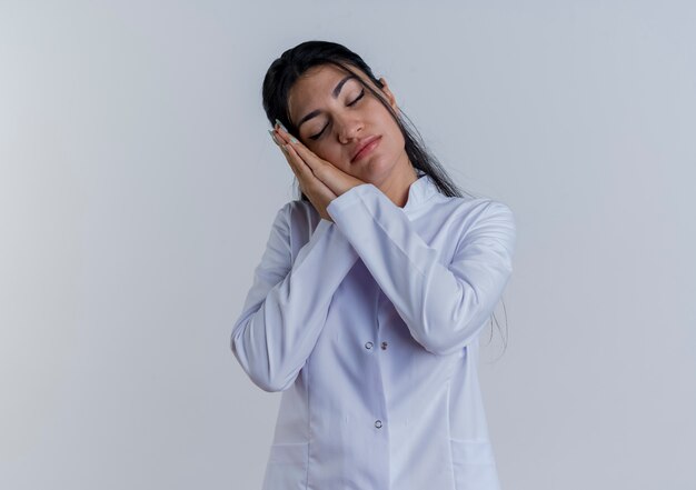 Doctora joven cansada vistiendo bata médica haciendo gesto de sueño con los ojos cerrados aislados