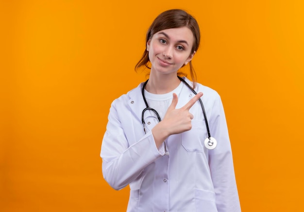 Doctora joven complacida con bata médica y un estetoscopio apuntando hacia el lado derecho en el espacio naranja aislado con espacio de copia