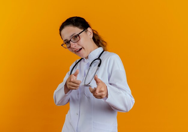 Doctora joven complacida con bata médica y estetoscopio con gafas mostrando gesto aislado